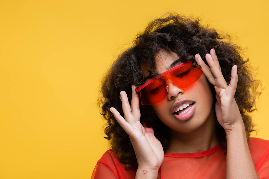 sensual mujer afroamericana con cabello ondulado ajustando gafas de sol de moda aisladas en amarillo - Foto, imagen