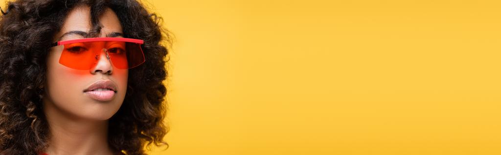 πορτρέτο της μελαχρινής Αφροαμερικανής με κόκκινα γυαλιά ηλίου κοιτάζοντας κάμερα απομονωμένη σε κίτρινο, banner - Φωτογραφία, εικόνα