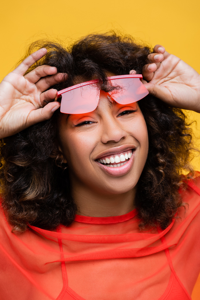 χαρούμενη Αφροαμερικανή γυναίκα που κρατάει κομψά γυαλιά ηλίου και κοιτάζει την κάμερα απομονωμένη στο κίτρινο - Φωτογραφία, εικόνα