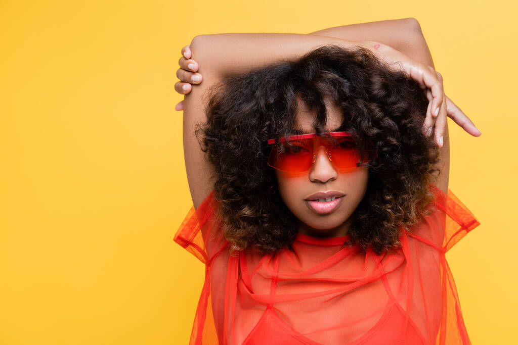 african american woman in red top and trendy sunglasses posing with hands above head isolated on yellow - Photo, Image