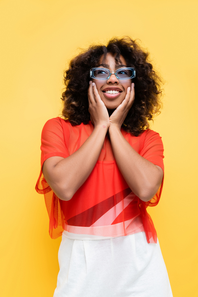 Африканська американка в модному одязі і сонцезахисних окулярах торкається обличчя і дивиться геть ізольовано на жовтий - Фото, зображення