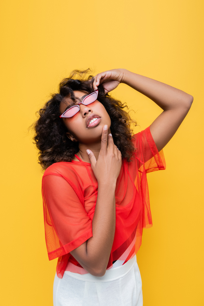 黄色で隔絶されたサングラスをかけながらあごや波状の髪に触れるアフリカ系アメリカ人女性 - 写真・画像