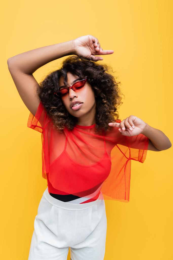 Afro-Amerikan kadın modayla gözlüklü ve güneş gözlüklü, sarı üzerine izole poz verirken parmaklarıyla işaret ediyor. - Fotoğraf, Görsel