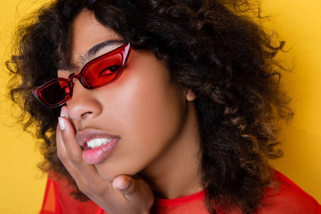 portrait of african american woman in red sunglasses touching face isolated on yellow - Photo, Image