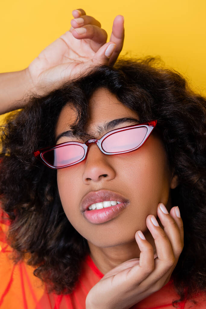 портрет кудрявой африканской американки в модных солнцезащитных очках, касающихся лица, изолированного на желтом - Фото, изображение