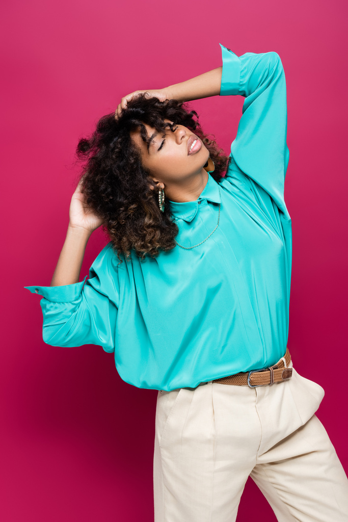 brunetta donna afroamericana con gli occhi chiusi toccare i capelli e posa isolata su rosa - Foto, immagini