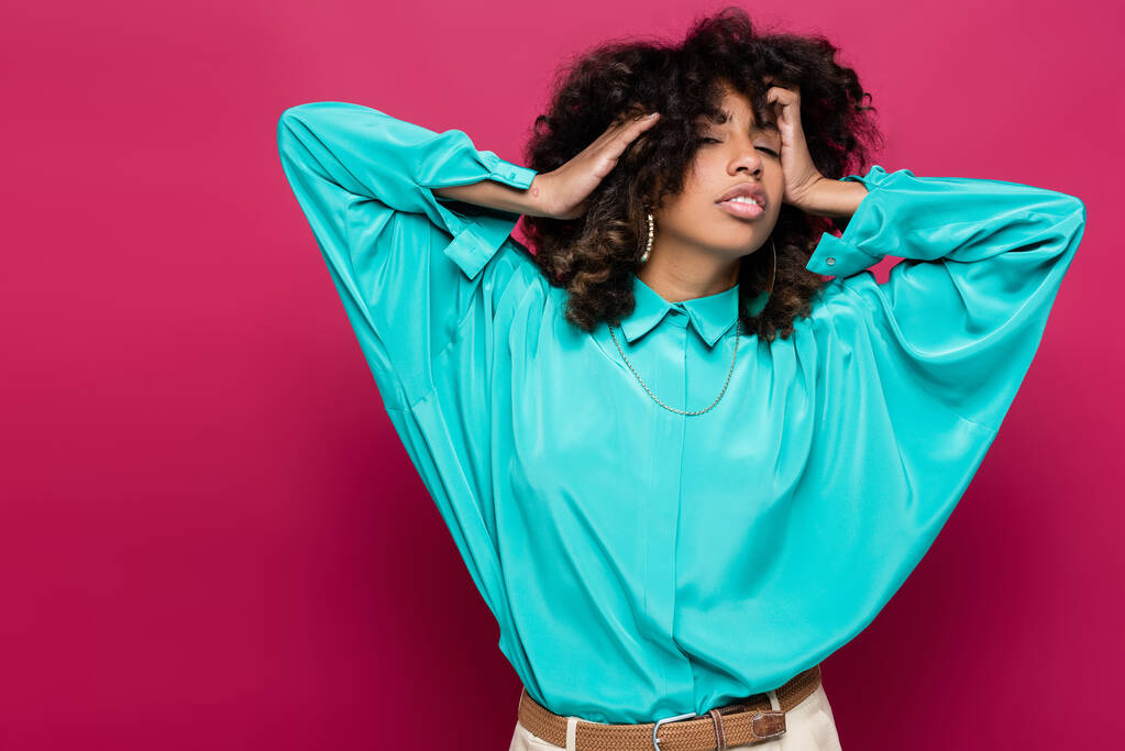 fashionable african american woman touching wavy hair while posing in blue blouse isolated on pink - Photo, Image