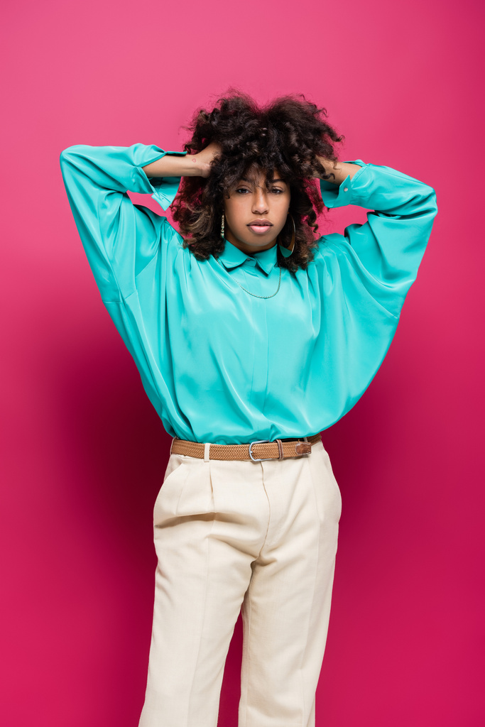 trendy african american woman in turquoise blouse touching curly hair isolated on pink - Photo, Image
