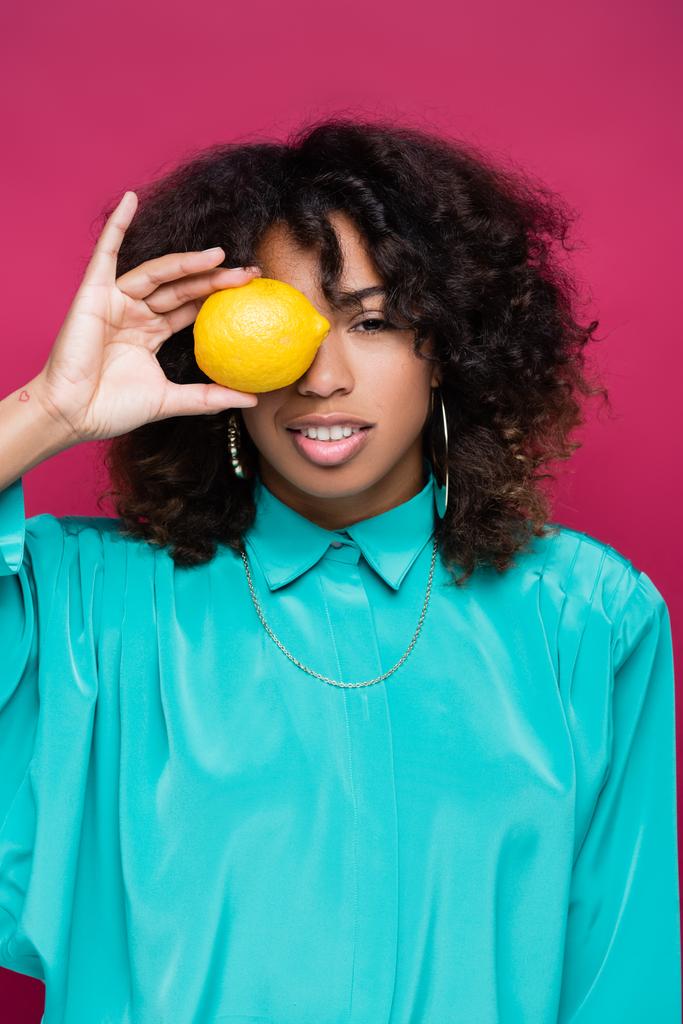 Африканская американка в голубой блузке закрывает глаза лимоном, изолированным на розовом - Фото, изображение