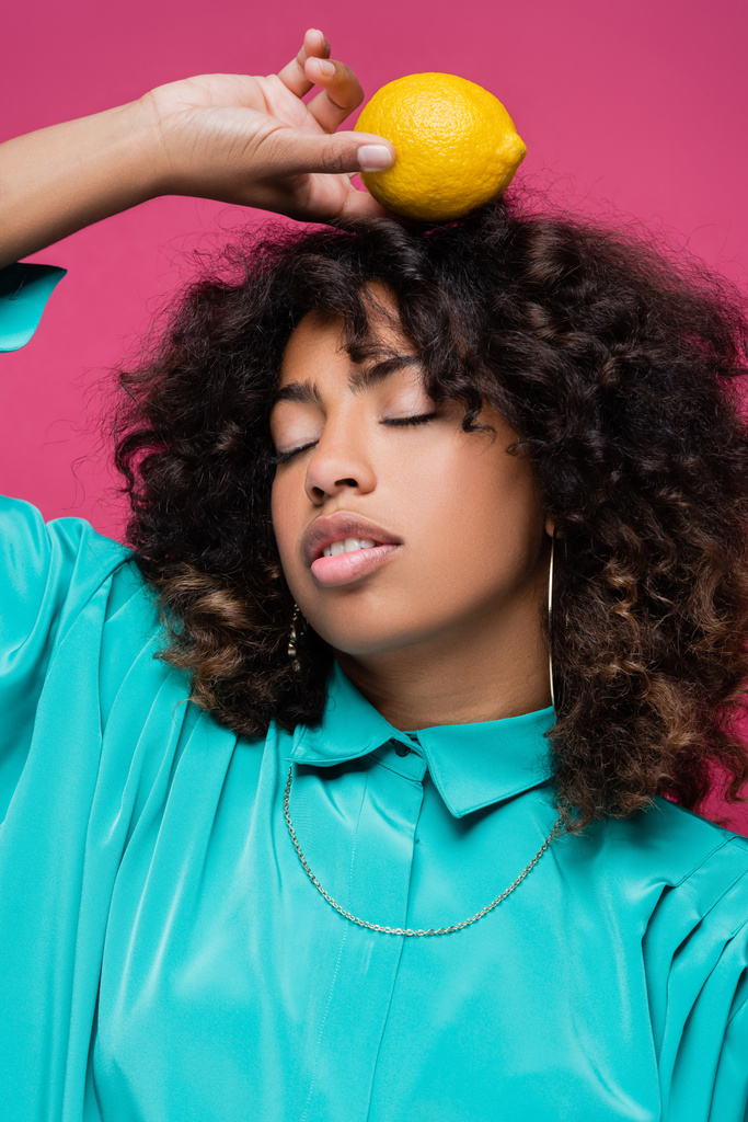 стильная африканская американка с закрытыми глазами, позирующая со спелым лимоном над головой, изолированным на розовом - Фото, изображение