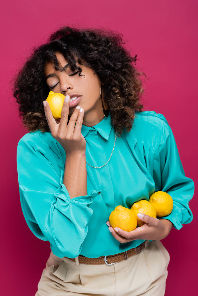 ピンク色に隔離された新鮮なレモンの匂いを嗅ぐ目を閉じたアフリカ系アメリカ人女性 - 写真・画像