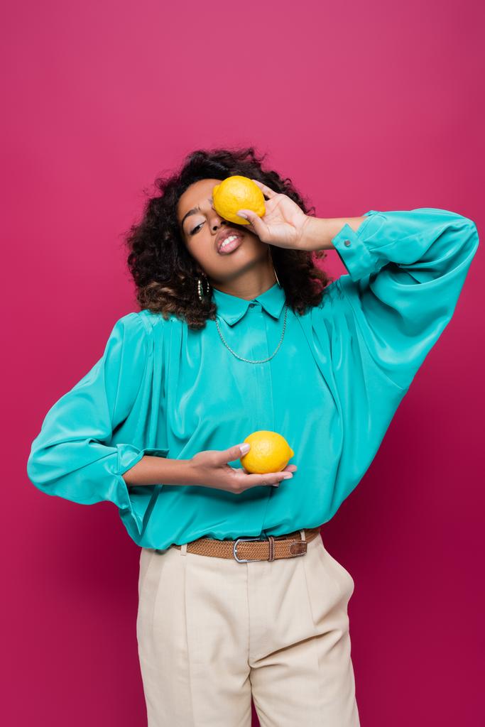 african american woman in blue blouse obscuring face with lemon isolated on pink - Photo, Image