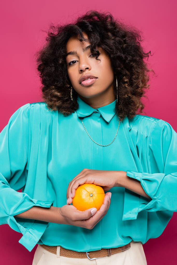 ピンクで隔離された熟したオレンジを保持している流行のシアンブラウスのアフリカ系アメリカ人女性 - 写真・画像