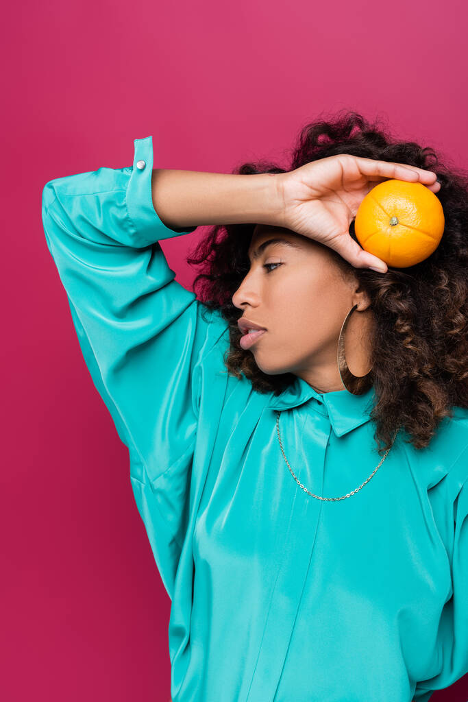 ricci donna afroamericana in posa con arancio maturo isolato su rosa - Foto, immagini