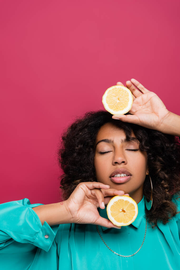 mujer afroamericana con los ojos cerrados sosteniendo mitades de jugoso limón aislado en rosa - Foto, imagen