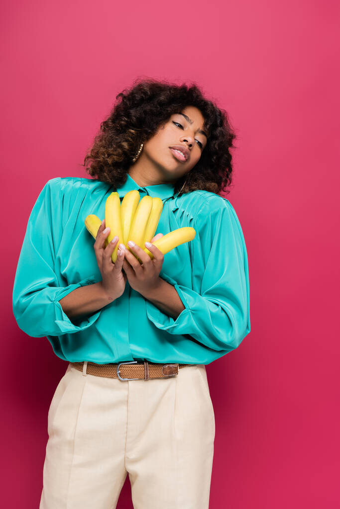  african american woman in stylish clothes holding ripe bananas isolated on pink - Photo, Image