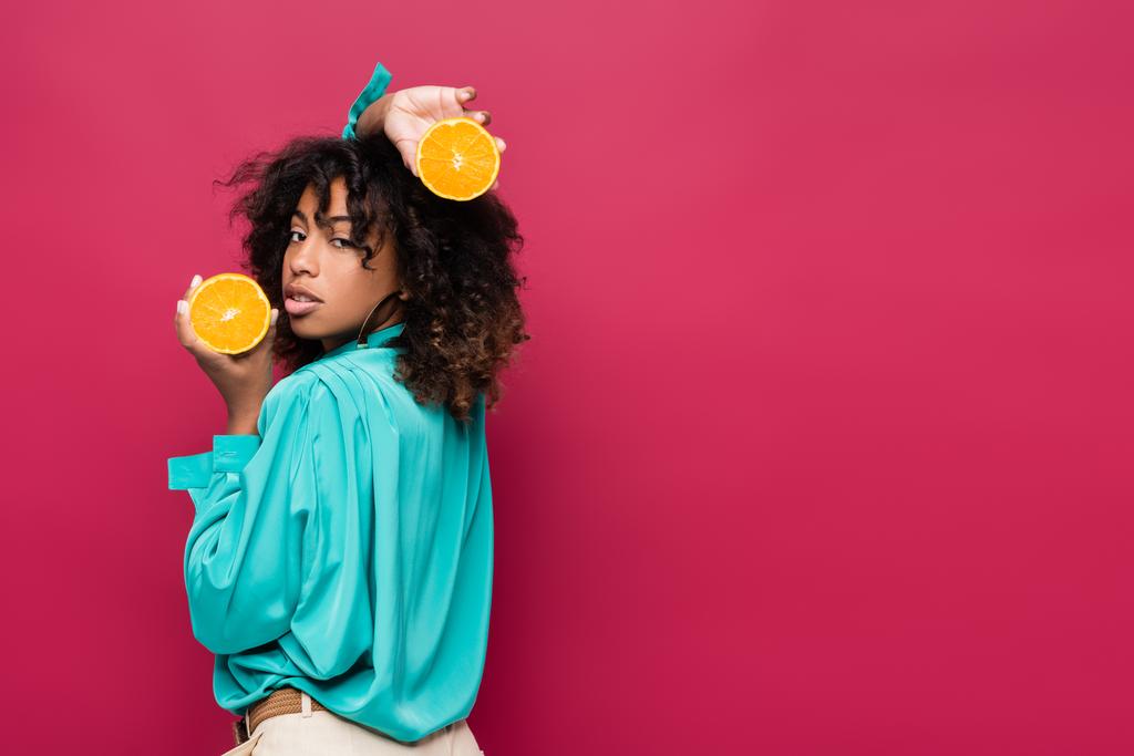 кудрявая африканская американка с половинками сочного апельсина, смотрящая на камеру, изолированную на розовом - Фото, изображение