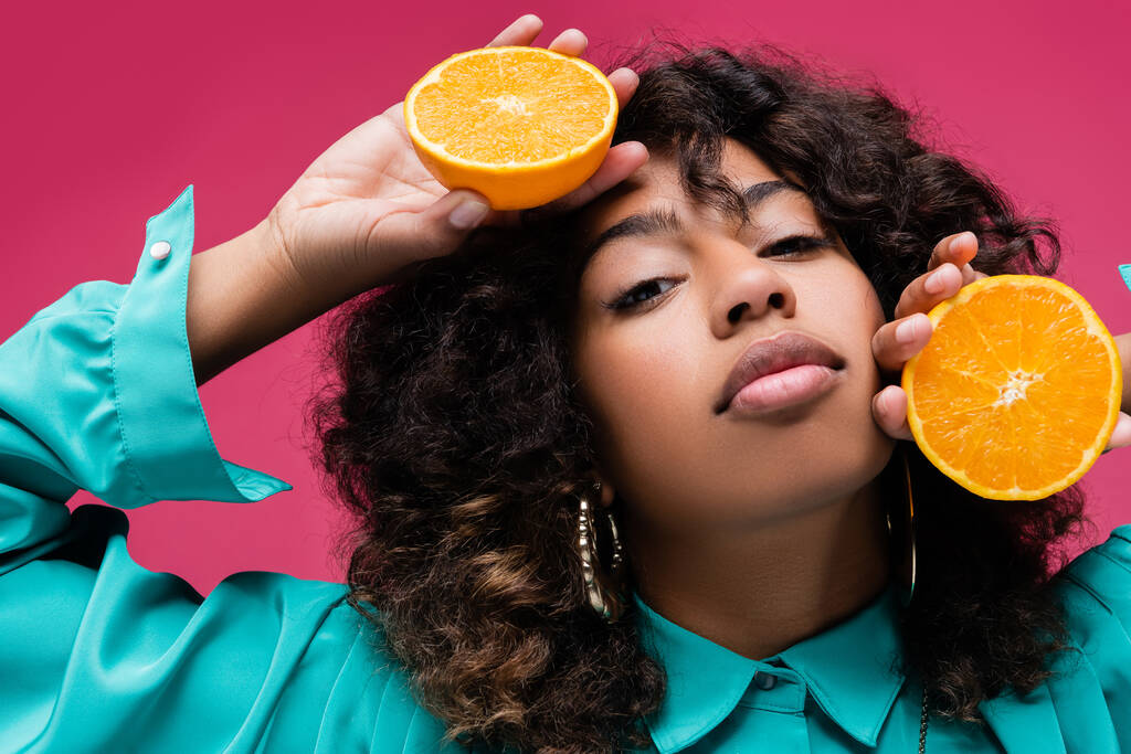 mujer afroamericana con el pelo ondulado sosteniendo mitades de naranja jugosa aislada en rosa - Foto, Imagen