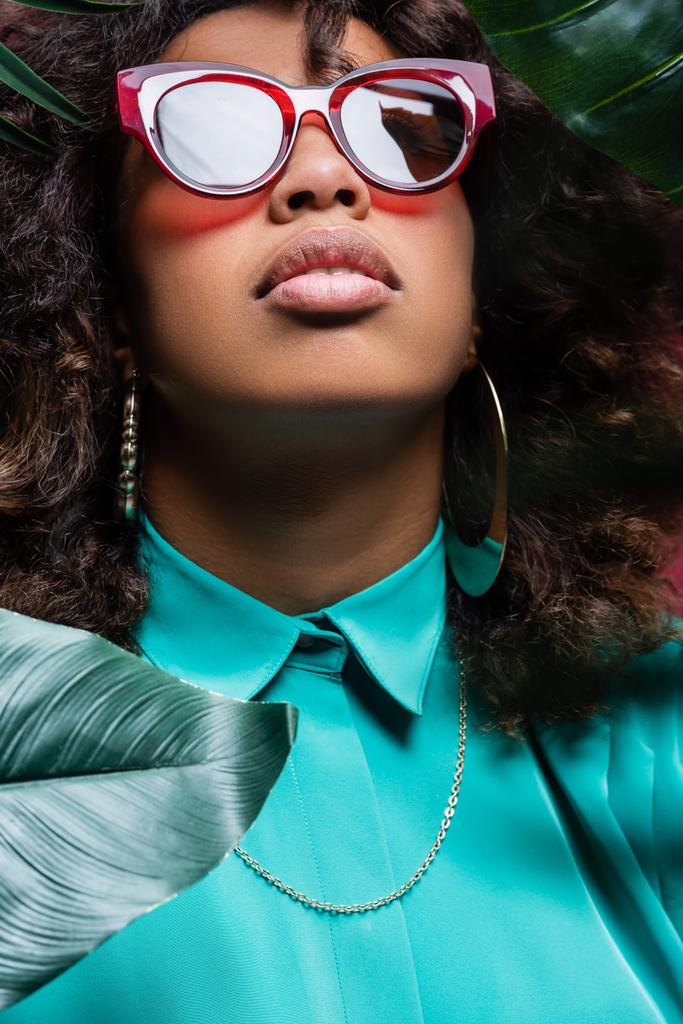 kudrnaté africké Američanky v náušnicích a slunečních brýlích v blízkosti zelených listů - Fotografie, Obrázek