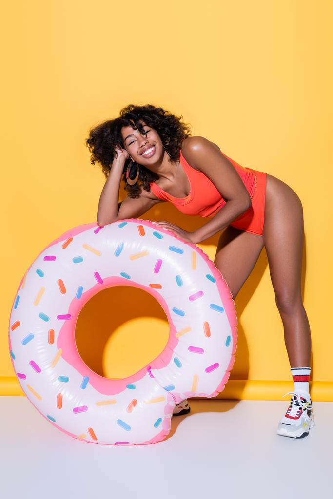 alegre mujer afroamericana en traje de baño sonriendo cerca de anillo de natación sobre fondo amarillo - Foto, imagen