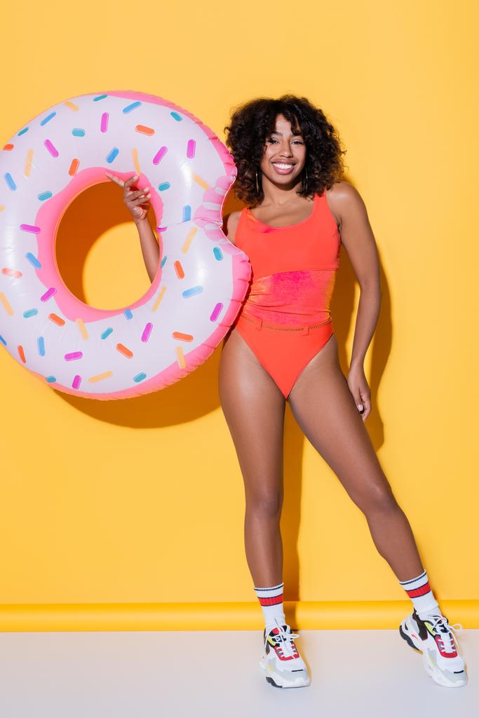 full length of happy african american woman in swimsuit and sneakers holding swim ring on yellow background - Photo, Image