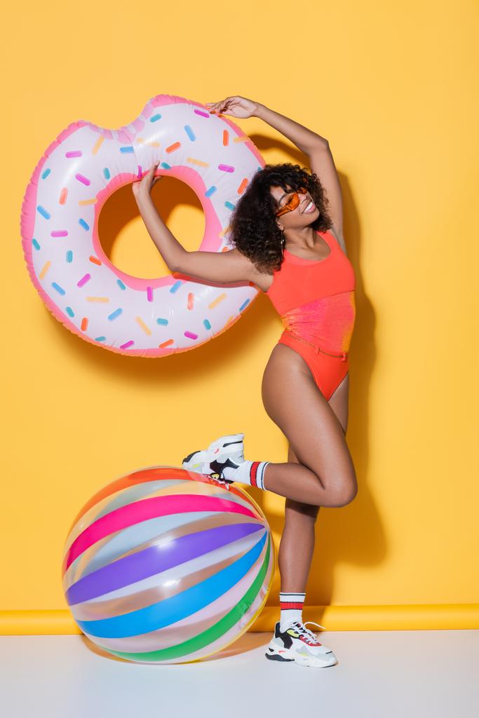 alegre mujer afroamericana en traje de baño posando con anillo de natación cerca de bola inflable sobre fondo amarillo - Foto, imagen