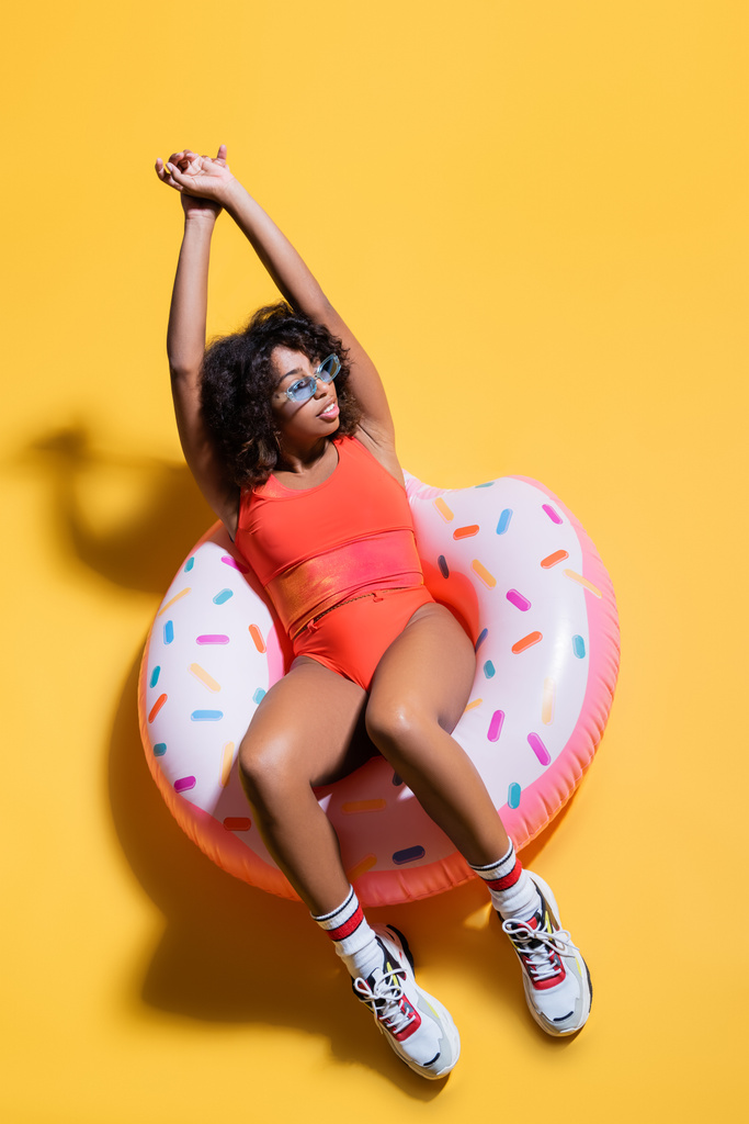 top view of african american woman in swimsuit and sneakers sitting on swim ring on yellow background - Photo, Image