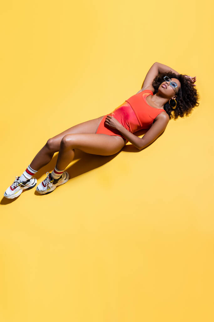 full length of african american woman in red swimsuit, sunglasses and sneakers on yellow background, top view - Photo, Image