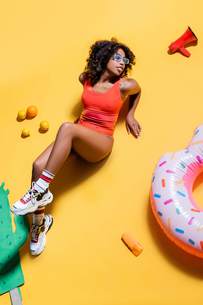 vista ad alto angolo della donna afro-americana in costume da bagno vicino all'anello di nuoto, frutta e altoparlante su sfondo giallo - Foto, immagini