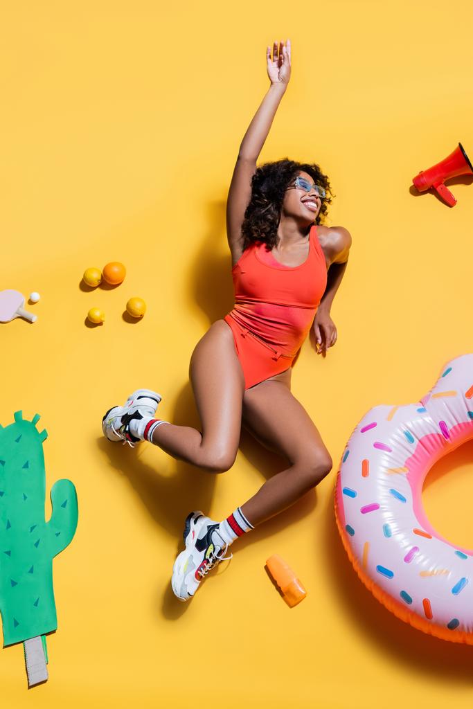mujer afroamericana feliz en ropa de playa cerca de frutas, anillo de natación y cactus cortados en papel sobre fondo amarillo - Foto, Imagen