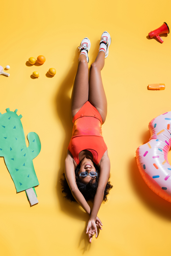 top view of african american woman in sunglasses and swimsuit lying near paper cut cactus and swim ring on yellow background - Photo, Image