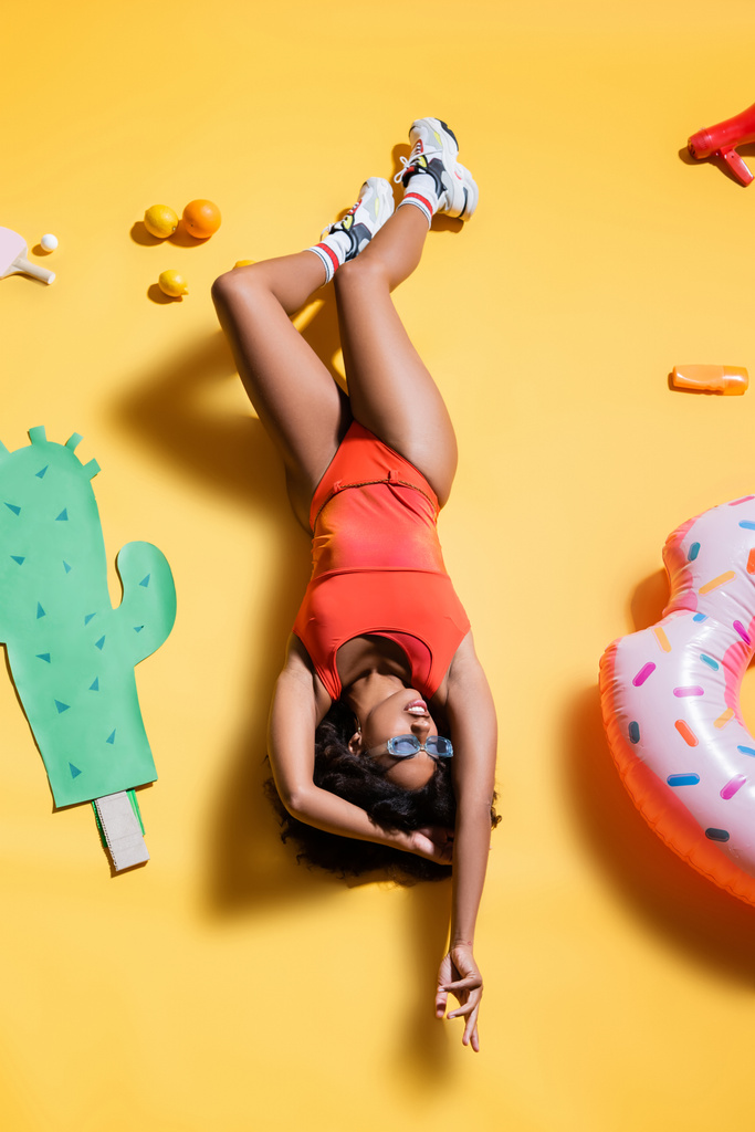 vue du dessus de la femme afro-américaine en maillot de bain relaxant près de l'anneau de natation et du cactus coupé en papier sur fond jaune - Photo, image