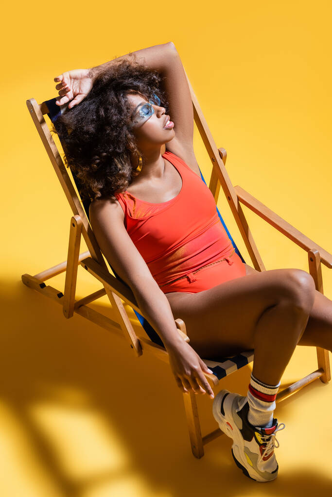 brunette afrikaanse amerikaanse vrouw in zonnebril en badpak rusten in ligstoel op gele achtergrond - Foto, afbeelding