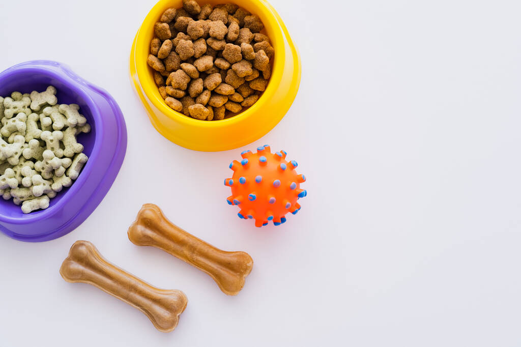 vista superior de galletas de mascotas en forma de hueso y comida seca en cuencos cerca de bola de goma aislada en blanco - Foto, imagen