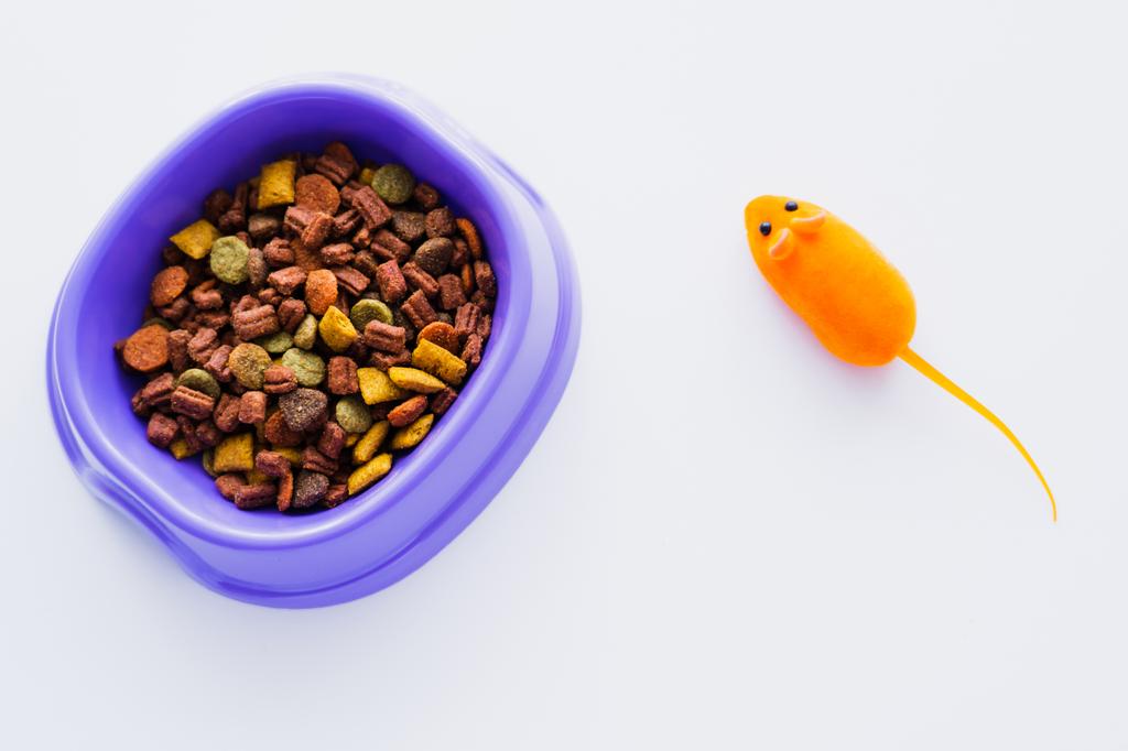 vista superior de tazón de plástico púrpura con comida seca para gatos cerca de ratón de juguete de goma aislado en blanco - Foto, Imagen