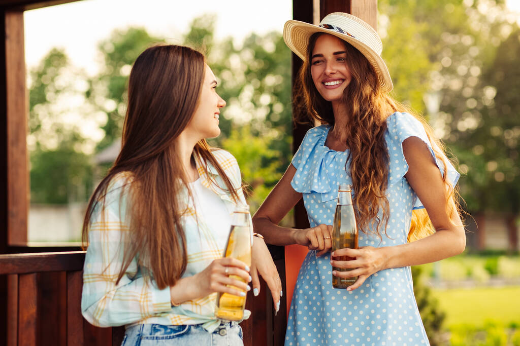Duas jovens mulheres bonitas e sorridentes em roupas de verão e chapéus. Mulheres despreocupadas fazem um piquenique na rua. Meninas positivas falando bebidas, ao ar livre no parque na natureza - Foto, Imagem