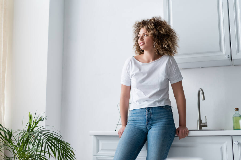 donna riccia in t-shirt bianca e jeans in piedi in cucina e guardando altrove - Foto, immagini
