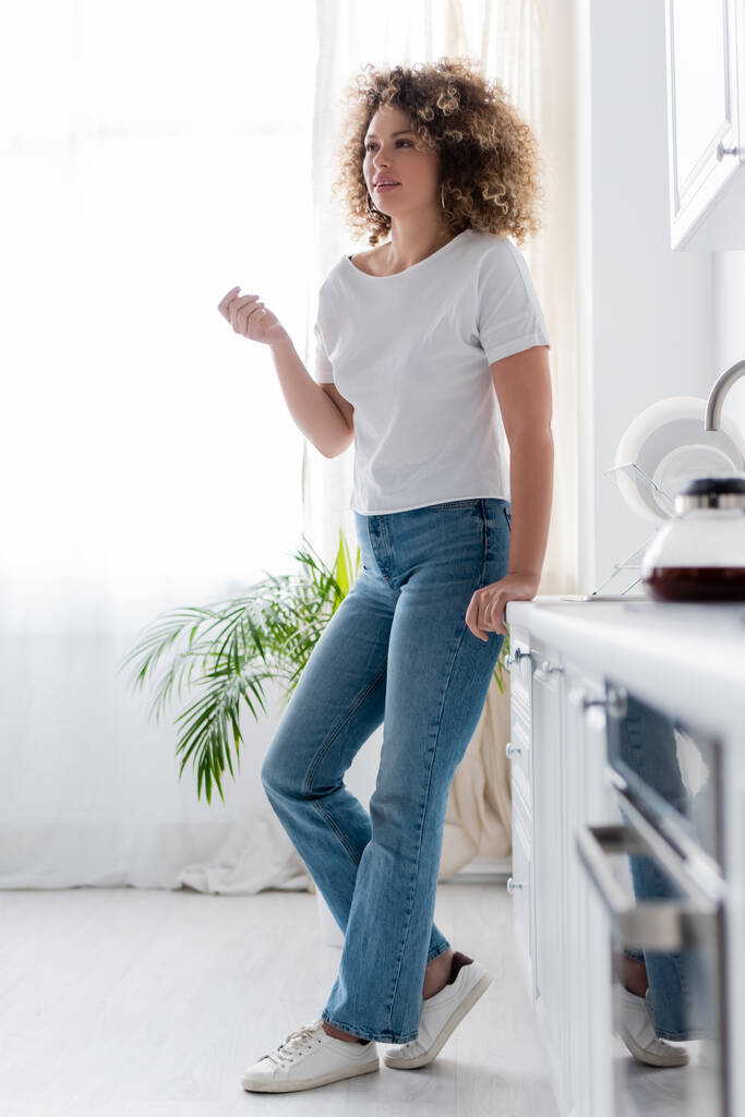 full length view of curly woman in jeans standing in kitchen on blurred foreground - Photo, Image