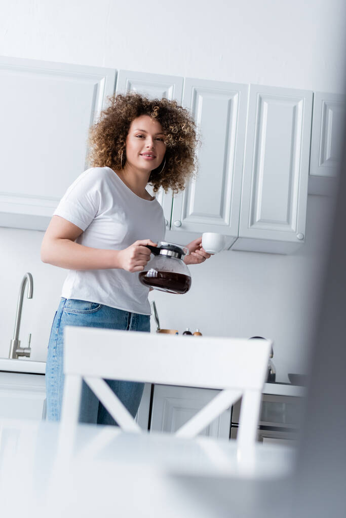 mulher encaracolado com panela de café e xícara sorrindo para a câmera na cozinha - Foto, Imagem