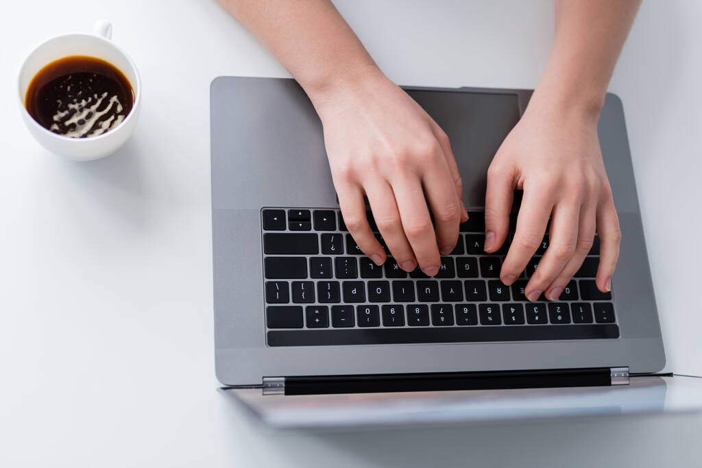 bovenaanzicht van bijgesneden vrouw typen op laptop in de buurt van kopje koffie - Foto, afbeelding