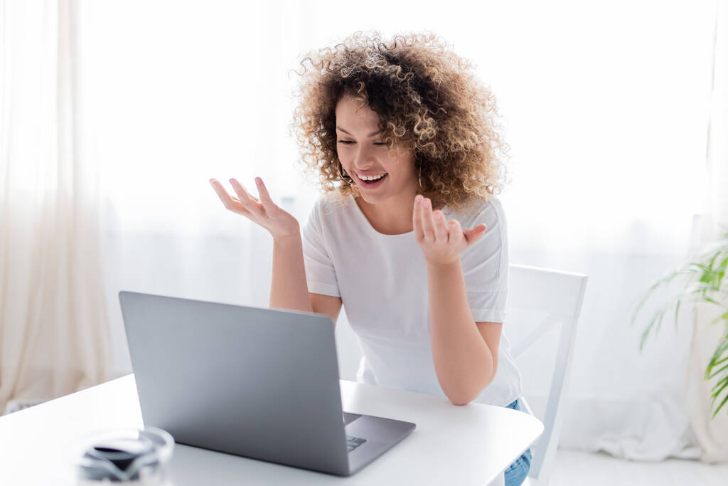 fröhliche Frau mit welligem Haar gestikuliert beim Videochat am heimischen Laptop - Foto, Bild