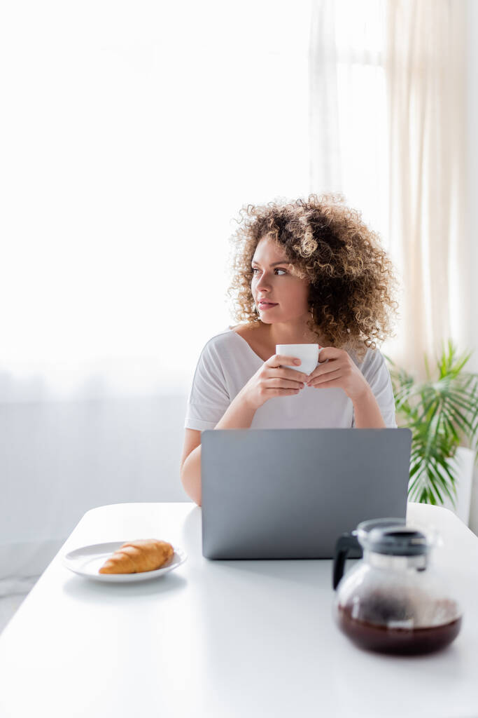 kręcone kobieta siedzi z filiżanką kawy w pobliżu laptopa i smaczne croissant w kuchni - Zdjęcie, obraz