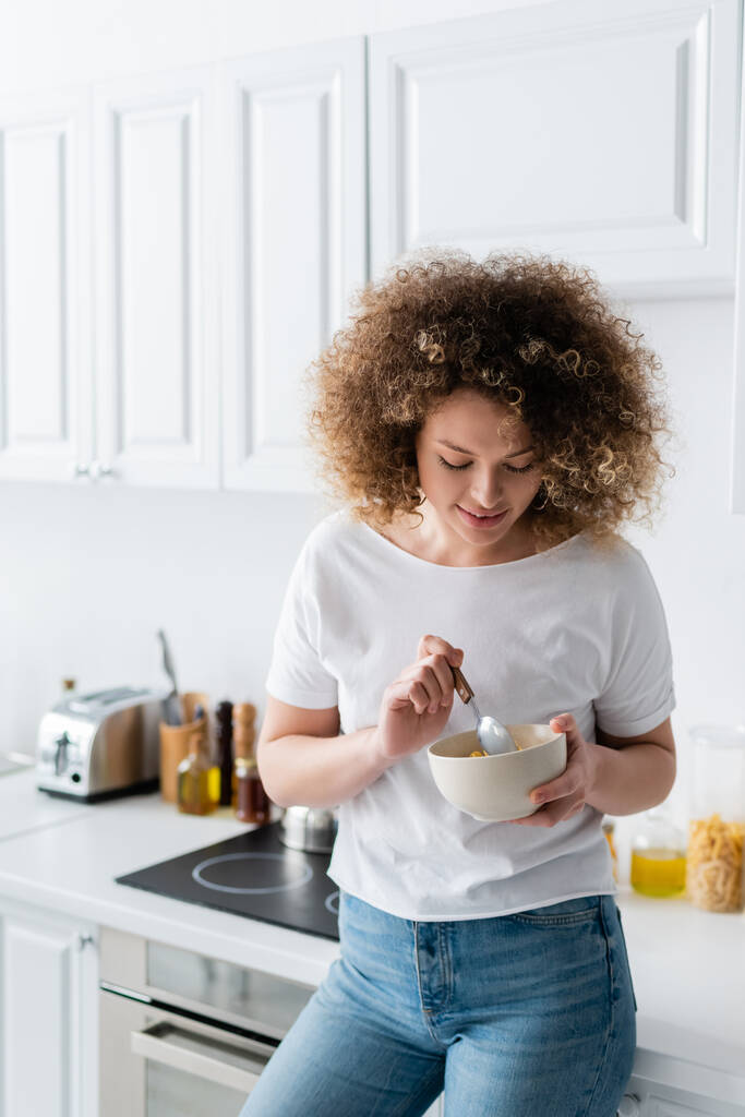mulher feliz com cabelo encaracolado segurando colher e tigela de saborosos flocos de milho na cozinha - Foto, Imagem