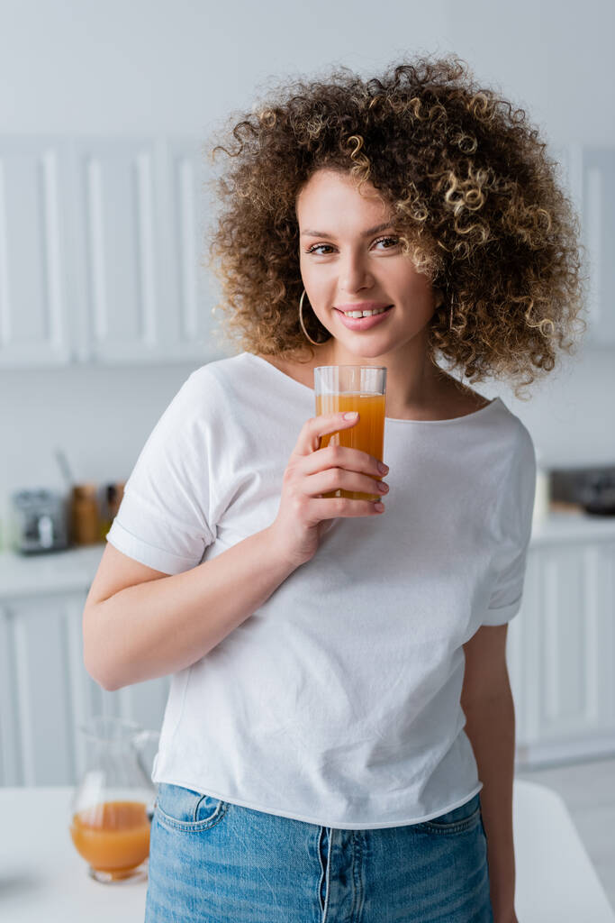 jong krullend vrouw in wit t-shirt met glas sinaasappelsap in keuken - Foto, afbeelding