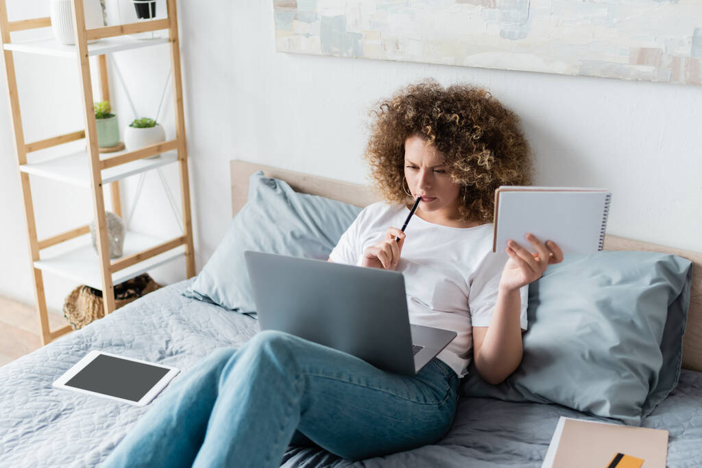 mulher pensativa com caneta e notebook trabalhando na cama perto de tablet digital - Foto, Imagem