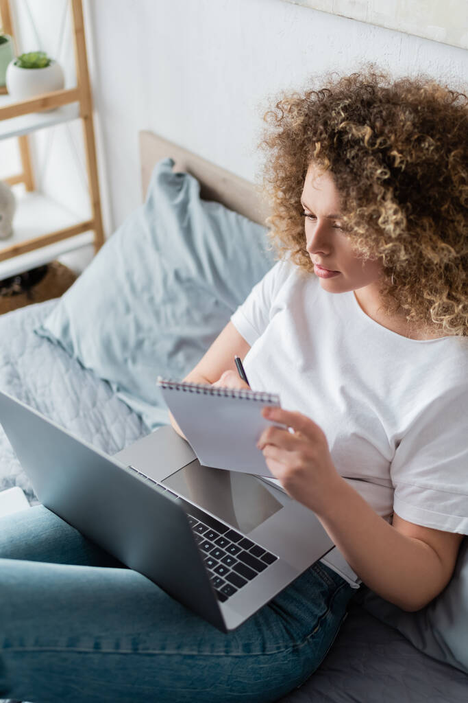 άποψη υψηλής γωνίας σγουρή γυναίκα κοιτάζοντας το laptop και γράφοντας στο σημειωματάριο στην κρεβατοκάμαρα - Φωτογραφία, εικόνα