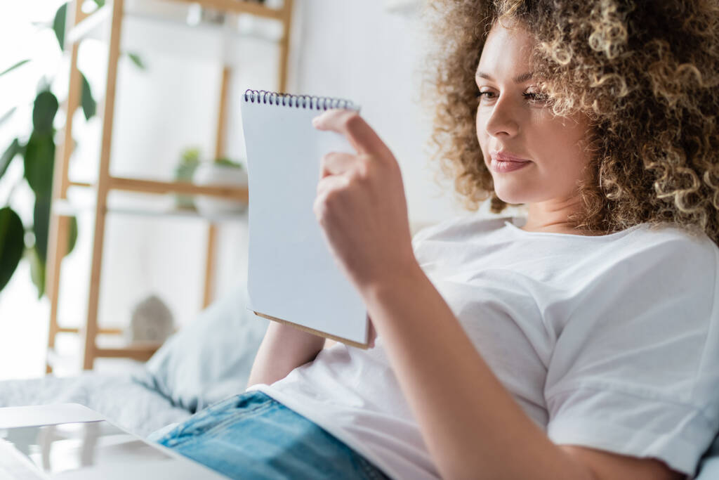 θετική γυναίκα με σγουρά μαλλιά κοιτάζοντας σημειωματάριο στο κρεβάτι στο σπίτι - Φωτογραφία, εικόνα