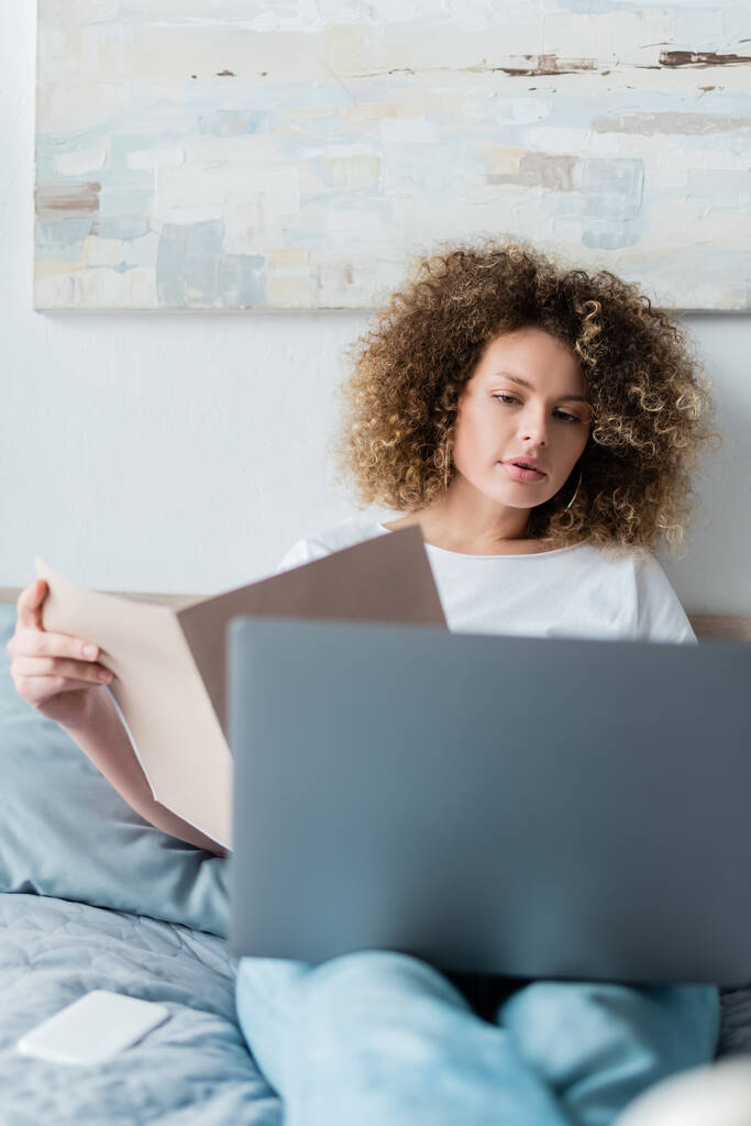 woman with wavy hair holding folder near laptop in bedroom - Photo, Image