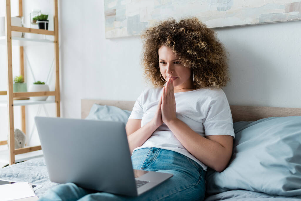 positive woman sitting with praying hands near laptop in bedroom - Photo, Image