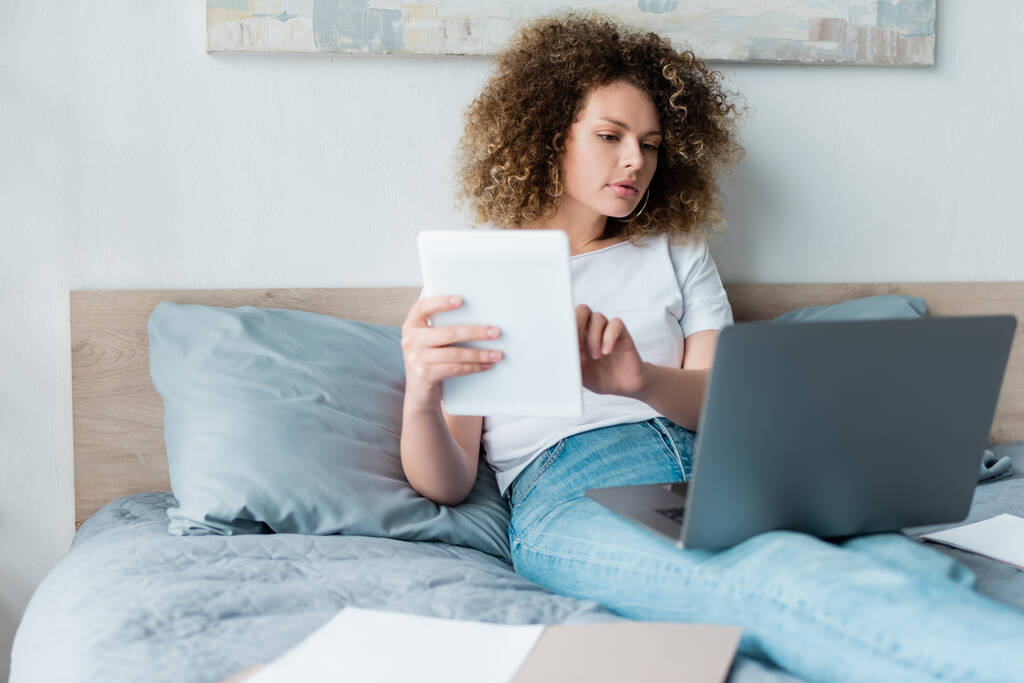 young curly woman working with digital tablet and laptop on bed at home - Photo, Image